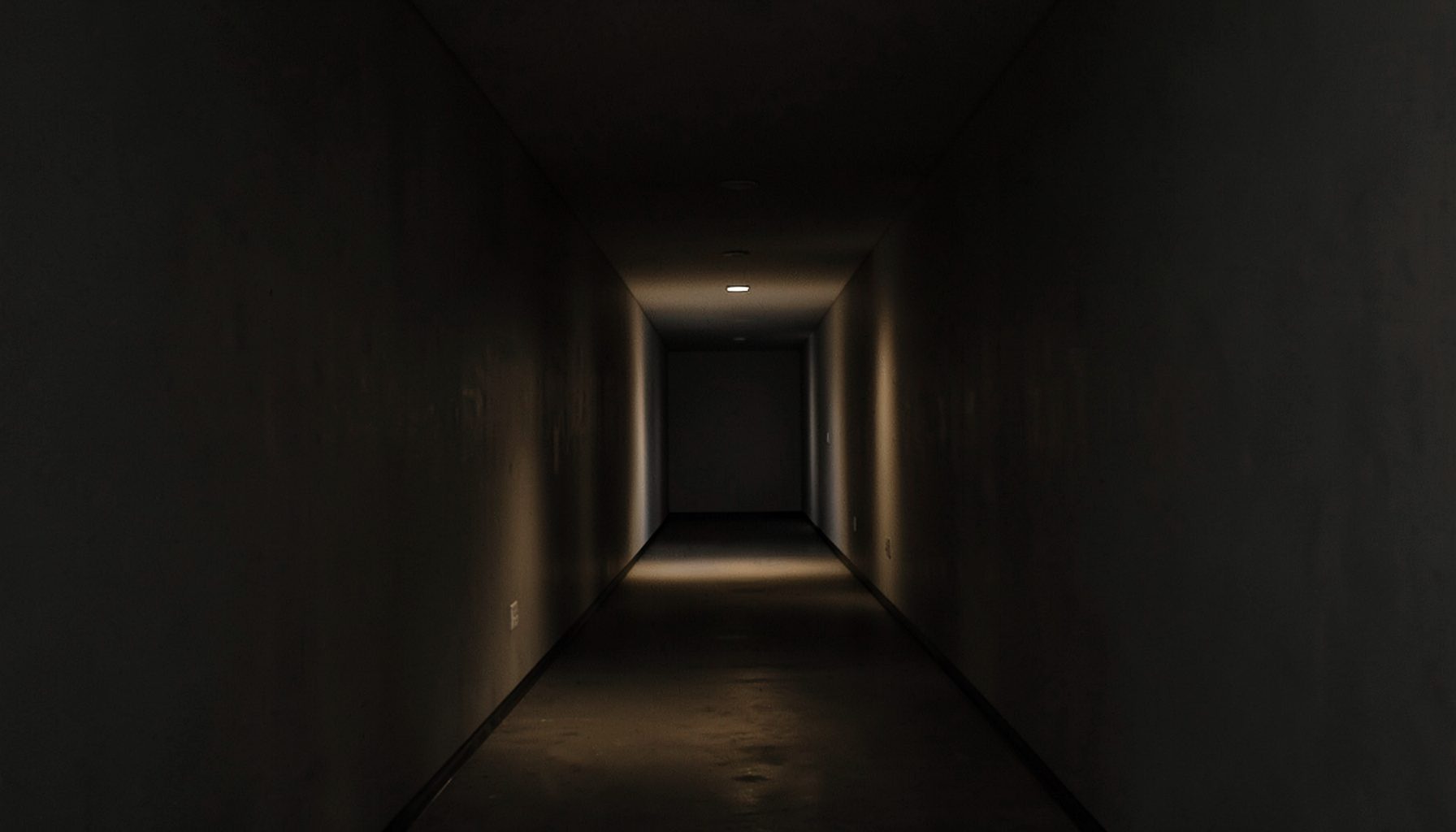 A shadowy corridor with a faint light at the far end, rendered in muted charcoal and amber tones
