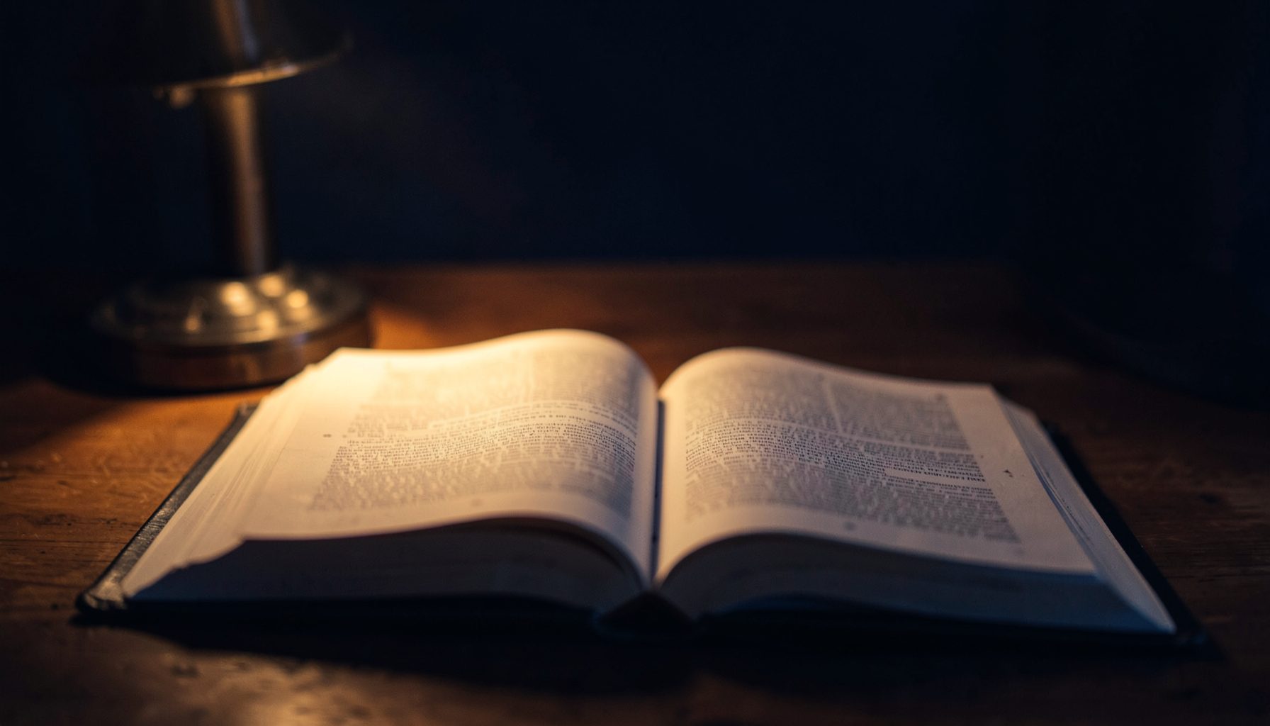 A dim, atmospheric view of an open Bible on a wooden desk with soft warm lamplight, deep navy and amber tones