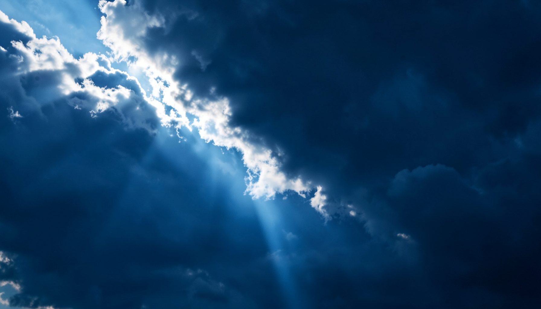 Clouds parting over a dark sky with a single bright beam of light breaking through, in deep blues and white