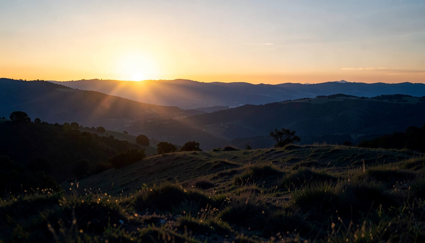 A wide, atmospheric landscape at dawn with soft golden light breaking over distant hills, a calm and hopeful mood in warm amber and deep blue tones