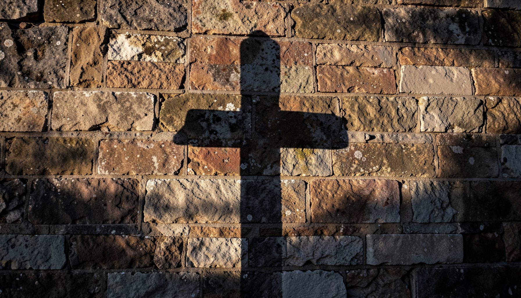 A weathered stone wall with a cross silhouette cast by afternoon light, in deep browns and warm grays